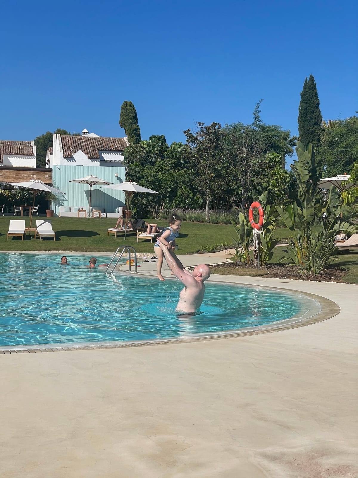 Parent lifting a toddler at the edge of La Zambra's family pool, with loungers, umbrellas and the hotel's blue-and-white Andalusian building behind.