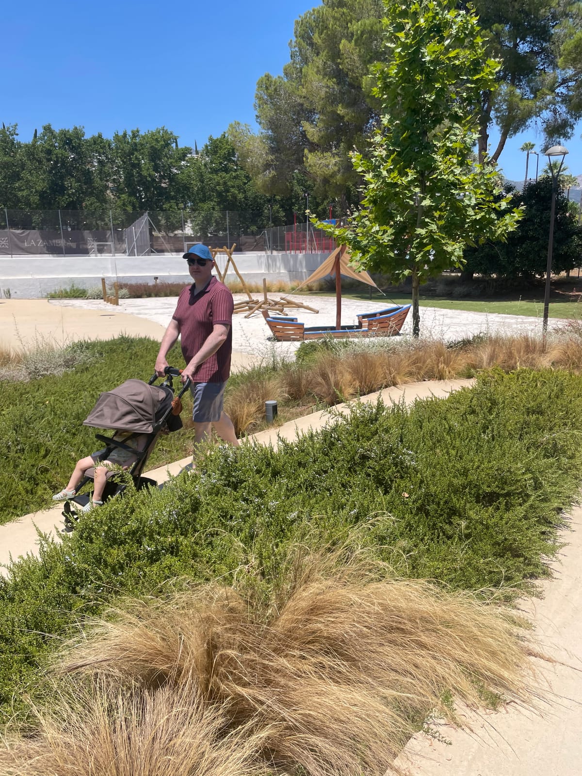 Parent pushing a stroller along a wide flat path through La Zambra's landscaped grounds, with the LA ZAMBRA sign and sports courts in the background.